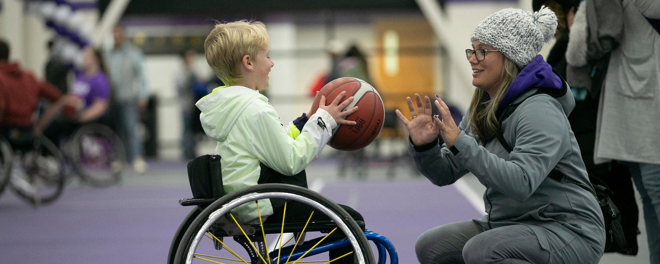 Boy in a wheelchair passing a basketball to a woman crouching in front of him