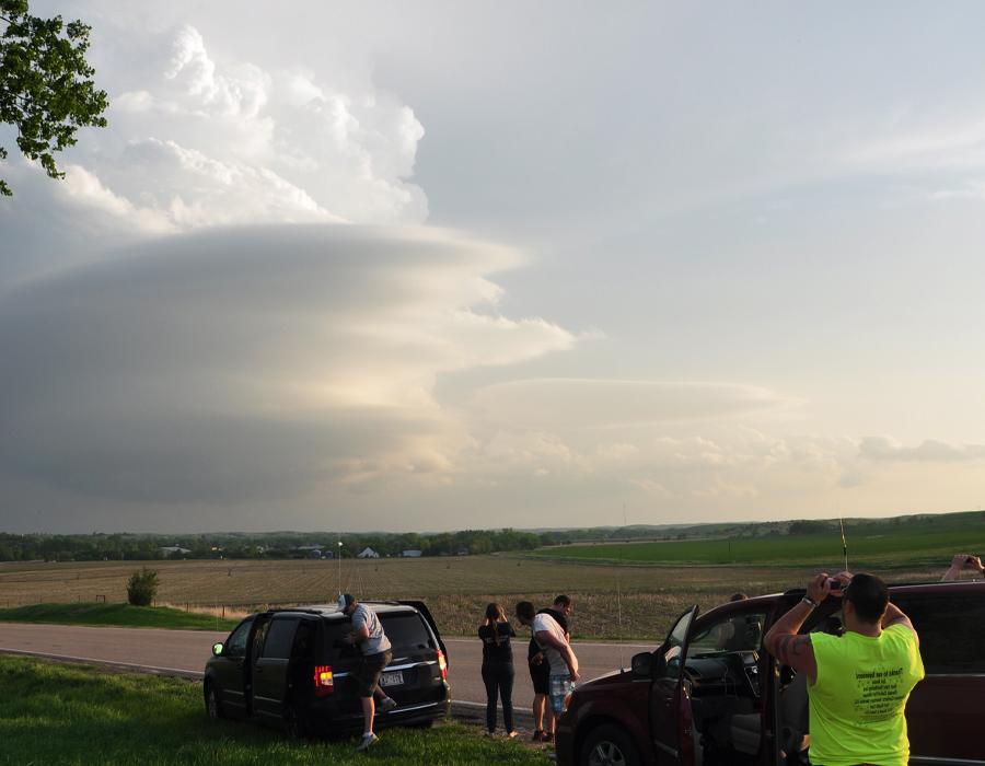 Students stand outside by vehicles, taking pictures of a stormy sky.